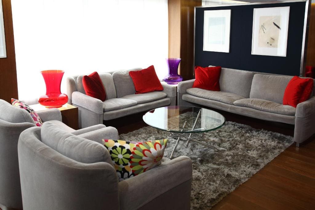 A modern living room with gray sofas, red and patterned pillows, a round glass coffee table, a shaggy gray rug, two red and one purple lamp, and framed artwork on the wall. Natural light fills the space.