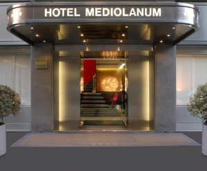 Entrance of Hotel Mediolanum with a modern glass door, two potted plants on each side, and a warmly lit interior visible, including a staircase, a red chair, and decorative lights inside.