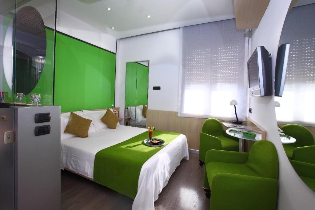Modern hotel room with a double bed, green accents on bedding and chairs, a wall-mounted TV, small glass table with two chairs, and large windows with blinds letting in natural light.