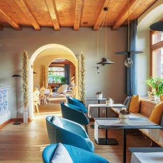 A cozy, sunlit cafe with wooden ceilings, blue chairs, white tables, and large windows. Plants and cushions decorate the space, and an archway leads to a brighter dining area in the background.