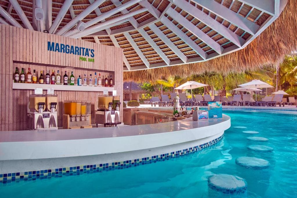 Swim-up pool bar called Margarita’s with bottles and drink dispensers on display, surrounded by stools in the water. Sunny outdoor setting with umbrellas and palm trees in the background.
