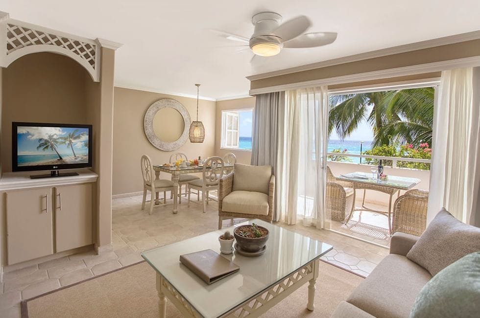 A bright, airy living room with beige furniture, a glass coffee table, TV, dining area, and sliding doors opening to a balcony with a sea view and palm trees. Sunlight fills the space, creating a relaxing atmosphere.