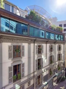 A sunlit building with French windows, balconies decorated with potted plants, and an upper glass terrace filled with greenery under a clear blue sky.