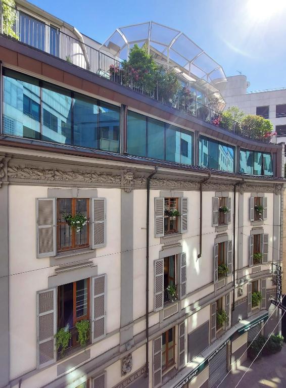 A sunlit building with French windows, balconies decorated with potted plants, and an upper glass terrace filled with greenery under a clear blue sky.