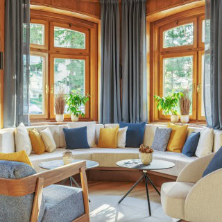 A cozy living room with large bay windows, blue and gray curtains, a curved sofa with blue, gray, and yellow cushions, plants on the windowsill, and round white coffee tables.