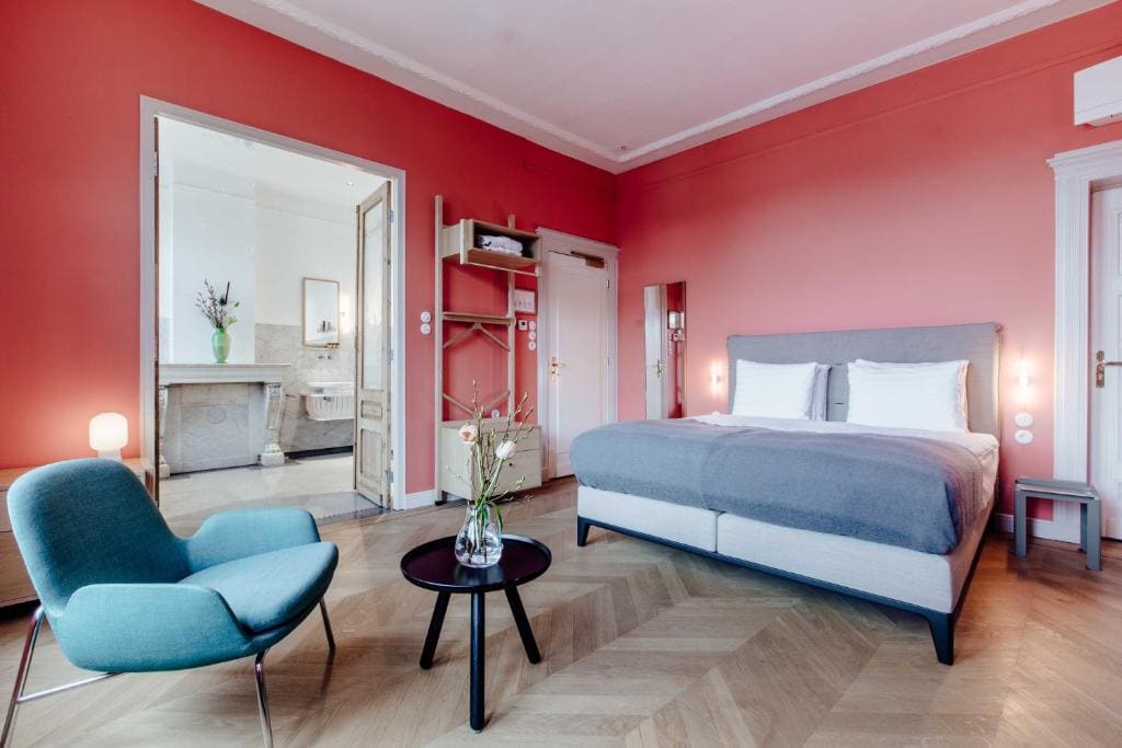 A stylish bedroom with coral pink walls, a large double bed with gray and white bedding, a blue armchair, a round table with a vase of flowers, wooden floors, and double doors leading to a bright bathroom.