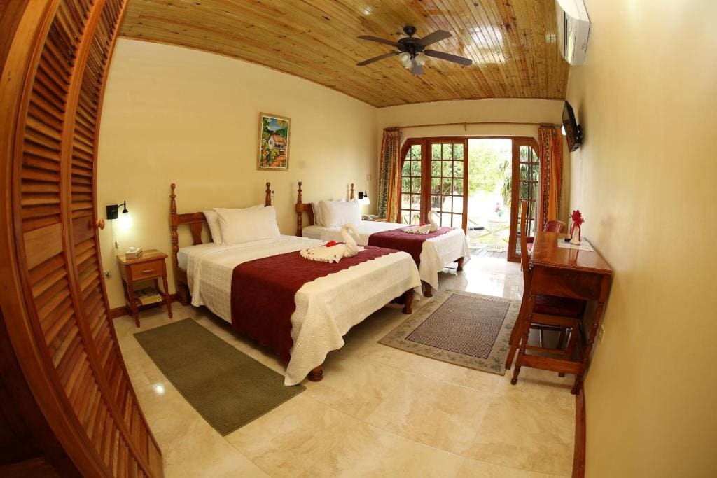 A cozy hotel room with two beds covered in white and burgundy bedding, a wooden ceiling, a desk, a ceiling fan, and large glass doors opening to a lush garden patio.