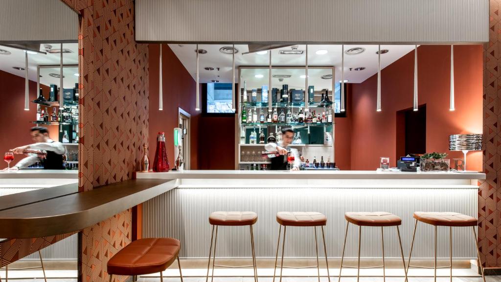 A modern bar with a white counter, four tan stools, and shelves lined with bottles. A bartender in a white shirt is moving behind the counter. The walls are red with patterned accents and soft overhead lighting.
