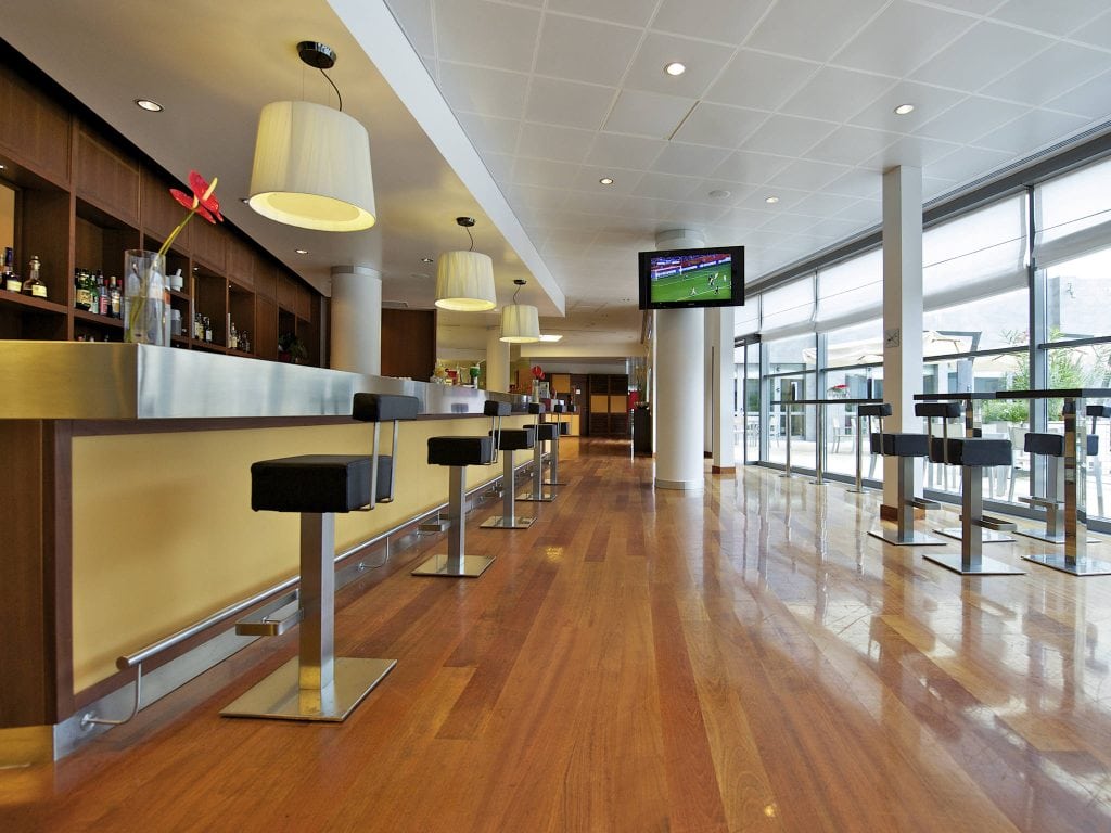 Modern bar with wooden floors, high stools, and a long counter. Large windows line the right side, letting in natural light. A TV is mounted from the ceiling, displaying a sports event. The space appears clean and spacious.