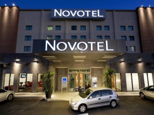A person stands by a parked silver car in front of a modern Novotel hotel entrance at dusk, with the building’s large illuminated signs visible above.