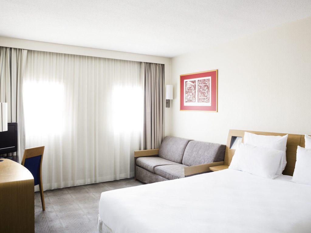 A bright, modern hotel room with a large bed, a gray sofa, a desk with a chair, and two windows covered by sheer curtains. A red-framed picture hangs on the wall above the sofa.