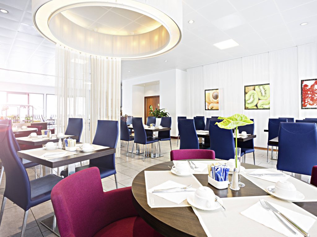 A modern, bright dining area with tables set for breakfast, featuring navy blue and maroon chairs, white dishware, placemats, and a large circular light fixture overhead. Artworks decorate the white walls.