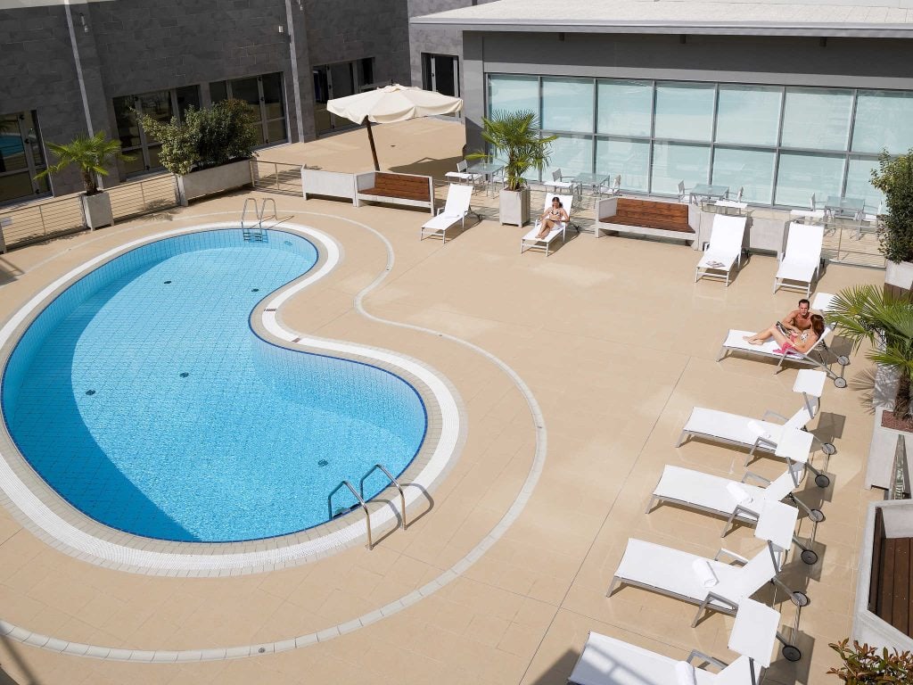 A kidney-shaped outdoor swimming pool is surrounded by white lounge chairs and palm trees. Two people are relaxing on loungers, and the area is bordered by modern buildings with large windows.