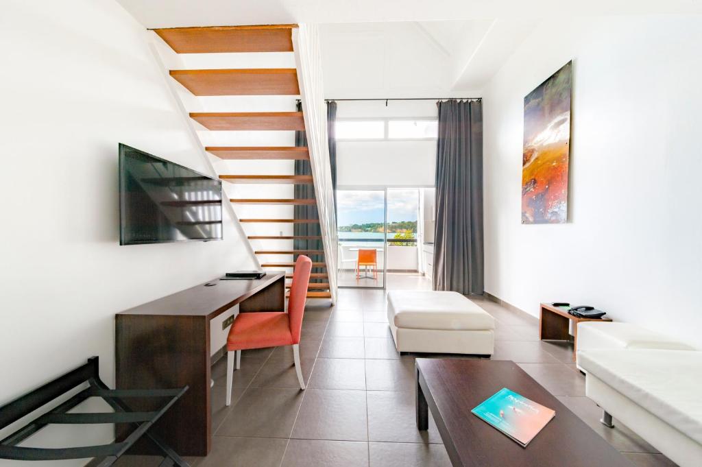 Modern, bright living space with a staircase, wall-mounted TV, desk, red chair, ottoman, coffee table, and balcony with a view of water and greenery. Large window lets in natural light; abstract art on the wall.