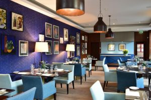A modern restaurant with blue and light teal chairs, dark wooden tables set for dining, a blue accent wall decorated with framed art, and warm pendant lights hanging from the ceiling.
