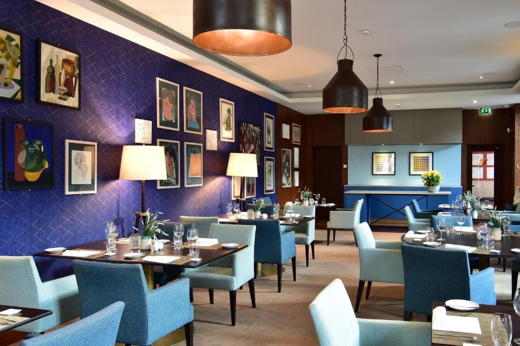 A modern restaurant with blue and light teal chairs, dark wooden tables set for dining, a blue accent wall decorated with framed art, and warm pendant lights hanging from the ceiling.