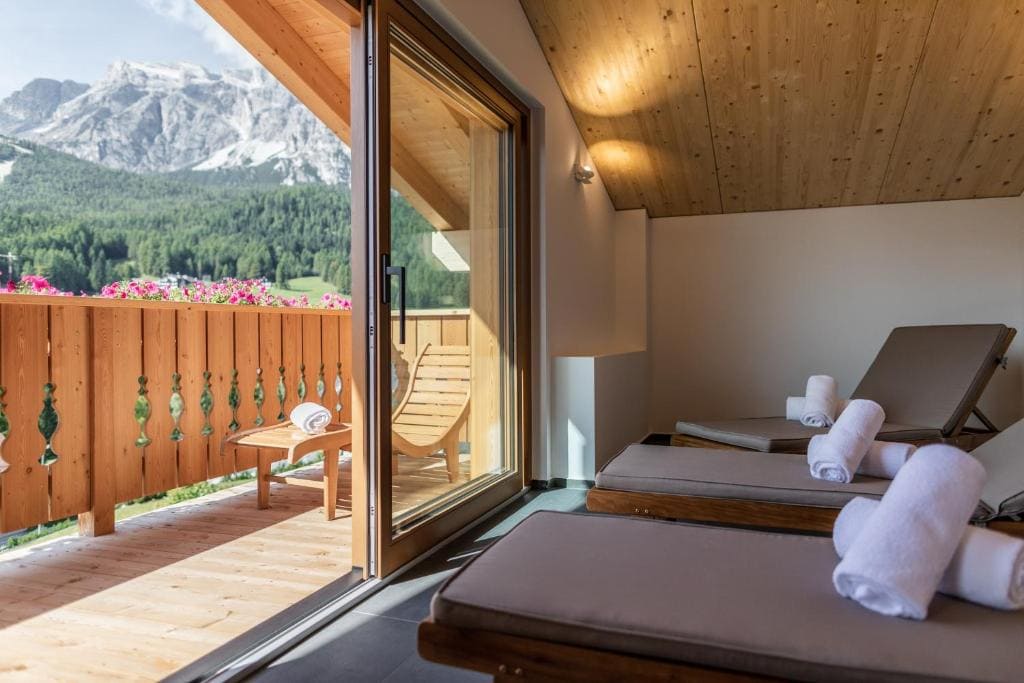 A cozy balcony with two loungers, rolled towels, wooden railing, and mountain views. Glass doors open to a wooden deck with a chair, table, and pink flowers, surrounded by alpine scenery.