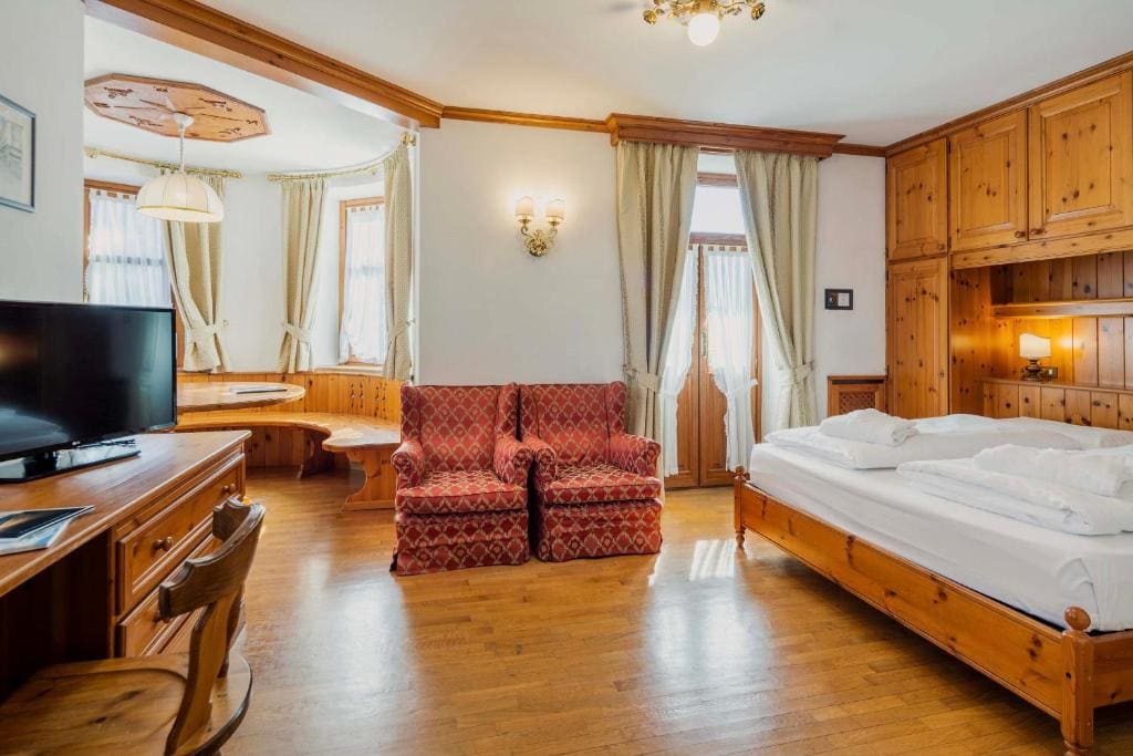 Bright hotel room with wooden furniture, a double bed, red patterned armchairs, a flat-screen TV on a desk, a round dining nook by bay windows, and large windows with beige curtains.