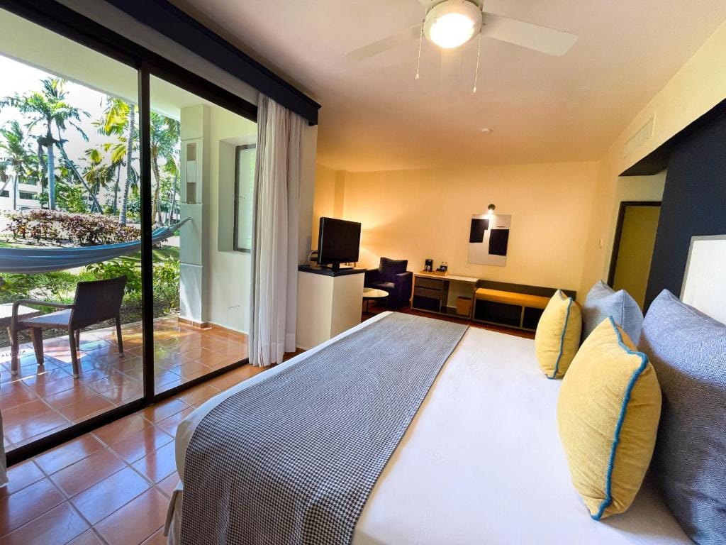A bright hotel room with a large bed, yellow and gray pillows, a TV, seating area, and sliding glass doors leading to a patio with chairs, table, and a hammock outside among greenery.