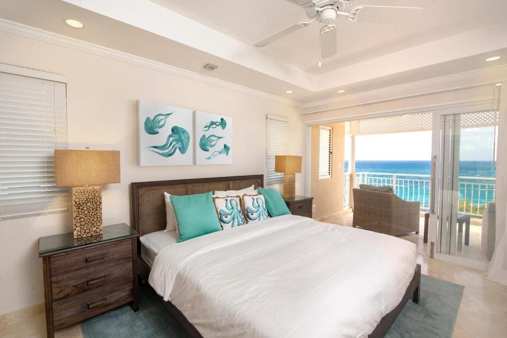 Bright bedroom with a large bed featuring teal pillows, two bedside tables with lamps, ocean-themed wall art, and sliding doors leading to a balcony with a view of the sea. Natural light fills the room.