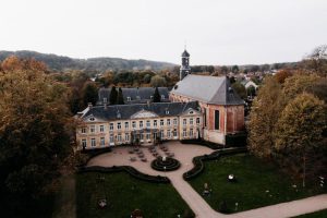 A large historic building with a central courtyard and round garden, surrounded by trees with autumn foliage. A church with a tall steeple is attached to the main structure; rolling hills and houses are visible in the background.