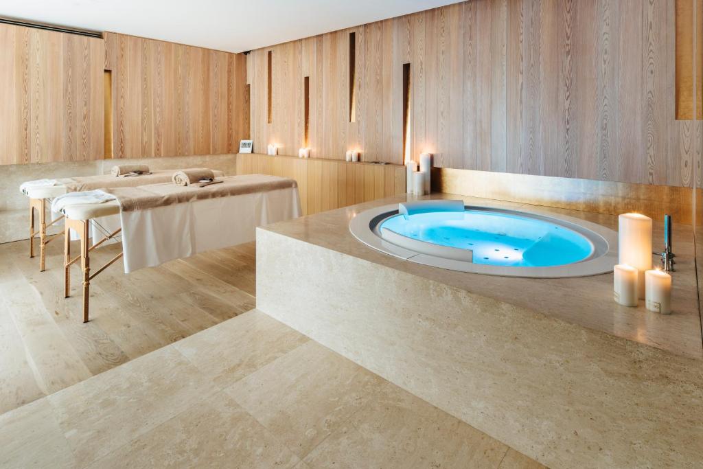 A modern spa room with two massage tables, beige stone flooring, wood-paneled walls, a round jacuzzi filled with blue water, and several lit candles creating a relaxing atmosphere.
