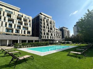Modern apartment buildings surround a rectangular outdoor swimming pool, with lounge chairs on green grass under a clear blue sky.