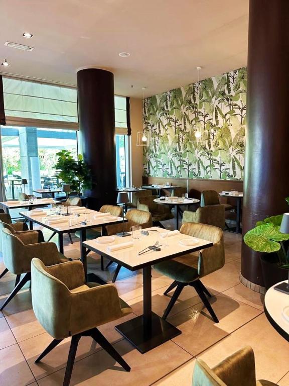 A modern restaurant dining area with green velvet chairs, white tables set for guests, large dark columns, potted plants, and a wall covered in tropical leaf-patterned wallpaper. Natural light enters from large windows.