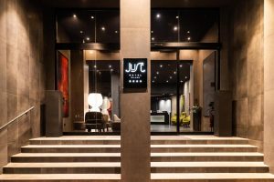 A well-lit hotel entrance with wide steps leading to glass doors. Inside, modern decor includes chairs and a reception desk. A sign above the entrance displays “JUS Hotel Milano” with four stars underneath.