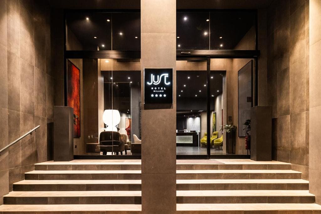 A well-lit hotel entrance with wide steps leading to glass doors. Inside, modern decor includes chairs and a reception desk. A sign above the entrance displays “JUS Hotel Milano” with four stars underneath.