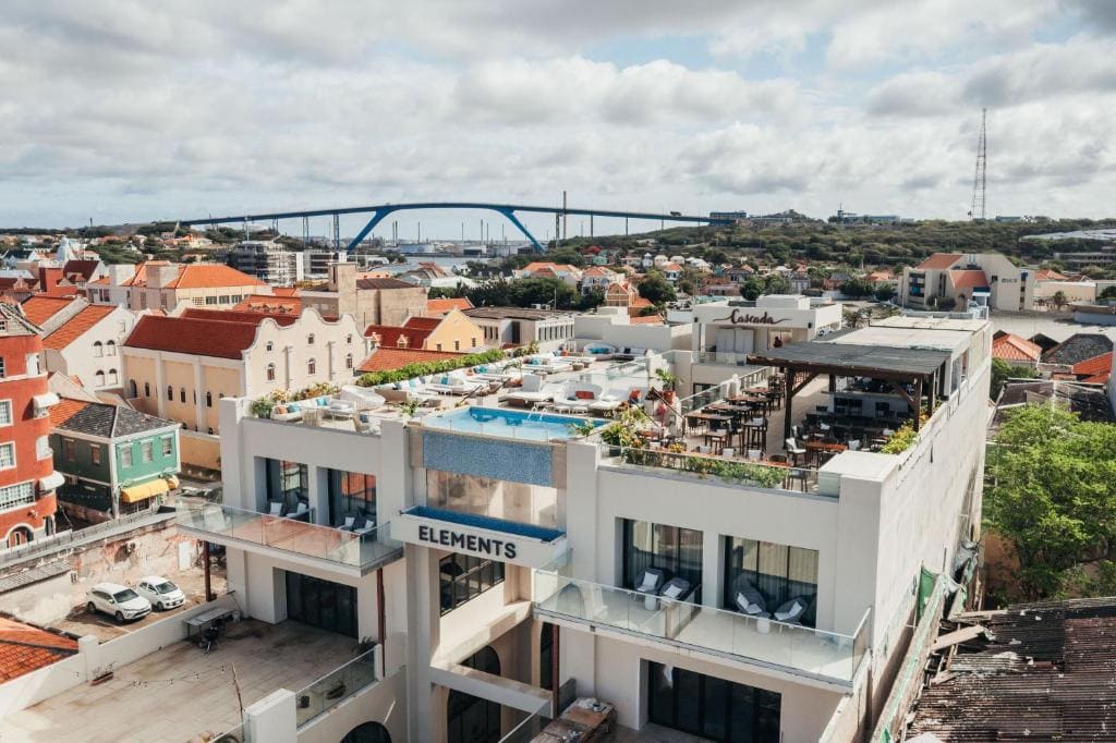 A rooftop pool and lounge area atop a modern building labeled ELEMENTS, surrounded by colorful houses, with a large bridge and cloudy sky in the background.
