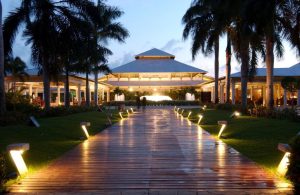 A wide wooden pathway lined with illuminated lights and palm trees leads to a large, open-air pavilion at dusk, with soft lighting highlighting the tropical resort ambiance.