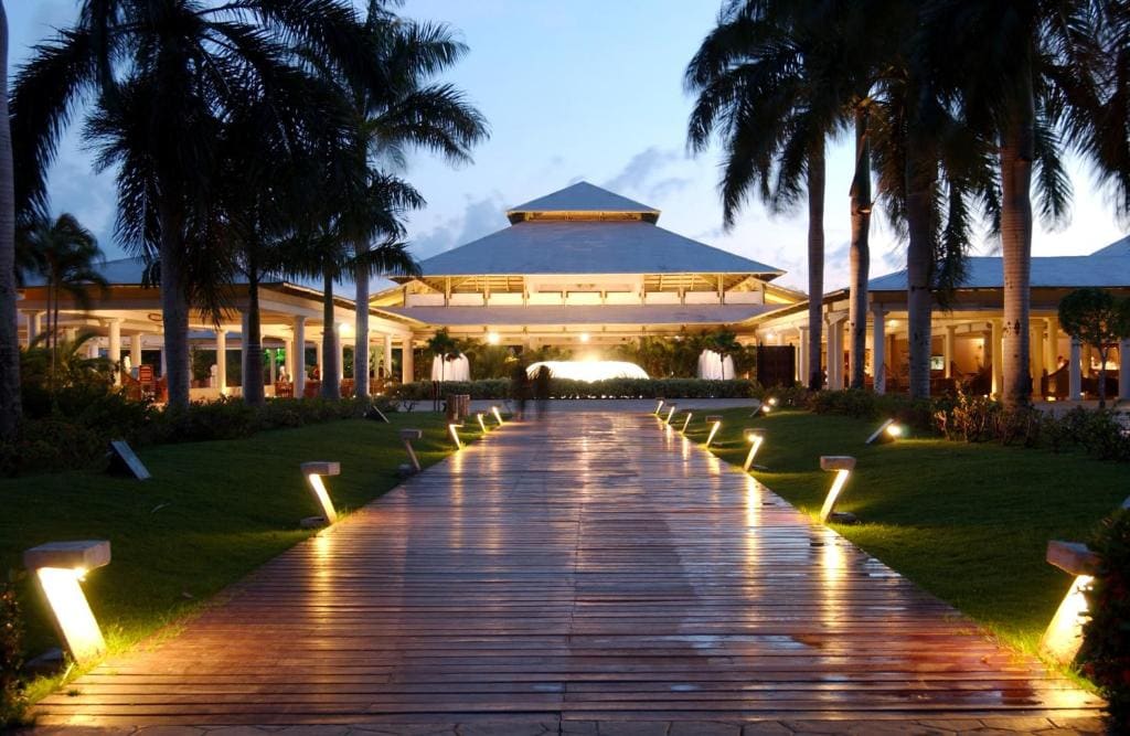 A wide wooden pathway lined with illuminated lights and palm trees leads to a large, open-air pavilion at dusk, with soft lighting highlighting the tropical resort ambiance.