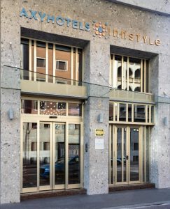 Entrance of AXYHOTELS InnStyle, featuring large glass doors and windows with beige frames, modern stone facade, and the hotel name in blue and orange lettering above the doors.