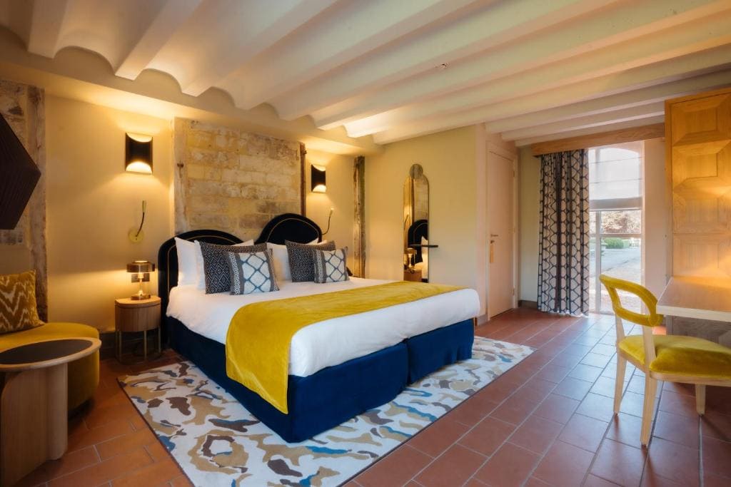A stylish hotel room with a large bed, yellow and blue accents, patterned rugs, wall lamps, a desk with a yellow chair, and sunlight streaming through a window with patterned curtains.
