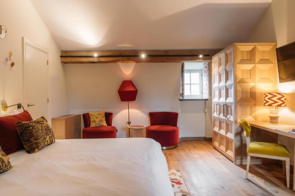 A cozy bedroom with a white bed, wooden floor, two red chairs, a hexagonal red lamp, a wooden wardrobe, a desk with a yellow chair, and a patterned lamp, with light coming from a small window.