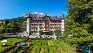 A grand hotel with red balconies and pointed towers stands behind a manicured garden with a small fountain, set against a backdrop of mountains and blue sky. Several cars are parked in front.