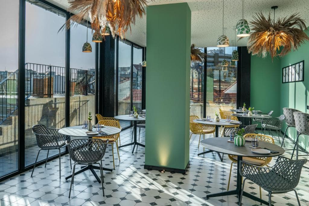 Modern restaurant interior with large windows, green walls, and black-and-white tile floor. Tables are set with plates and utensils, surrounded by woven chairs. Unique pendant lights with dried grasses hang from the ceiling.
