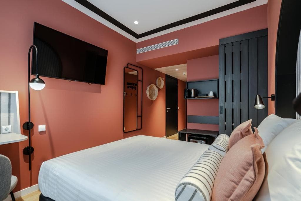Modern hotel room with a large bed, white bedding, and pink and gray accent pillows. The walls are terracotta-colored, and there is a wall-mounted TV, desk, and contemporary lighting fixtures.