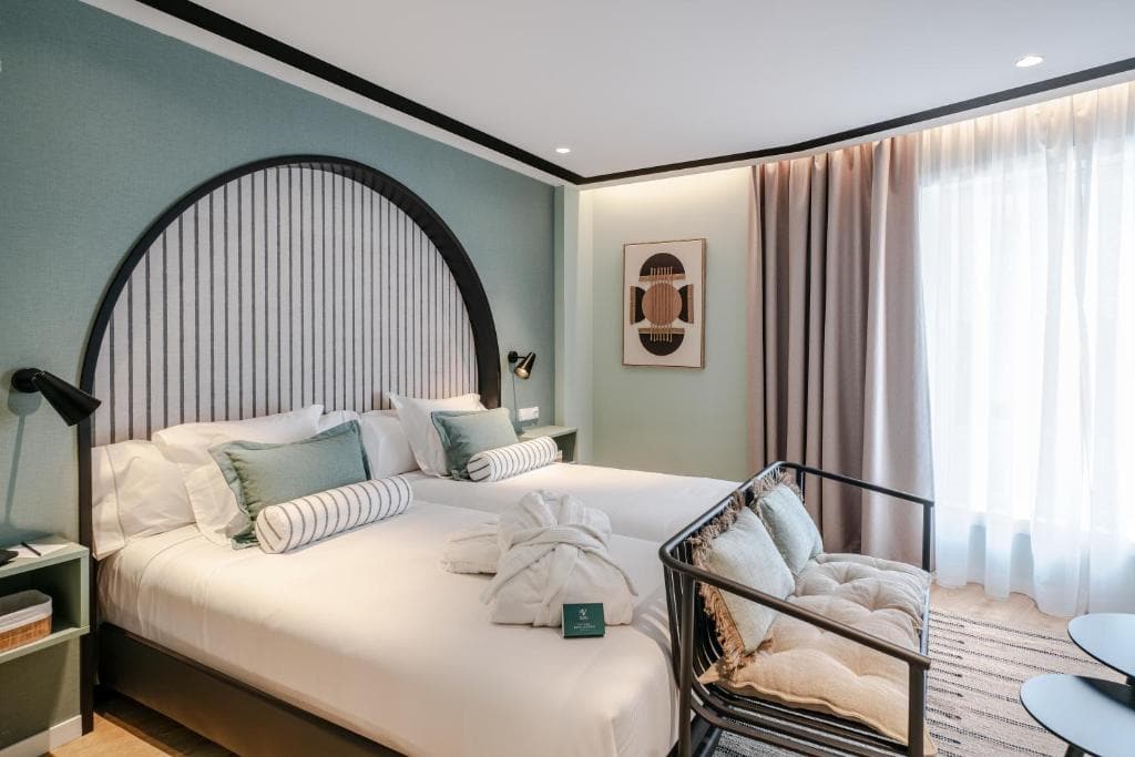 A modern hotel room with two twin beds side by side, decorative pillows, a bench at the foot of the beds, bathrobes, wall art, bedside lamps, light blue-green walls, and large curtained windows letting in natural light.