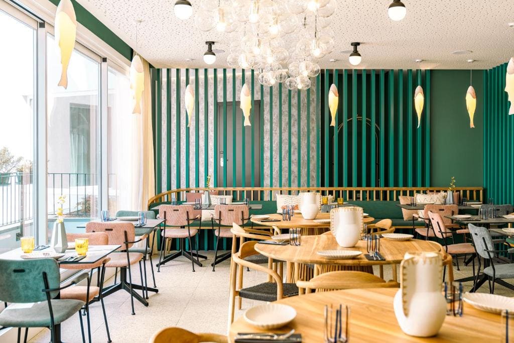 A modern restaurant with green vertical wall panels, light wooden tables, pastel chairs, large windows, decorative fish hanging from the ceiling, and a cluster of glass pendant lights.