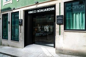 Entrance of Vincci Bonjardim Hotel with large glass doors, green-tiled exterior, and multiple signs, including one for a rooftop area. The street outside is paved with cobblestones.