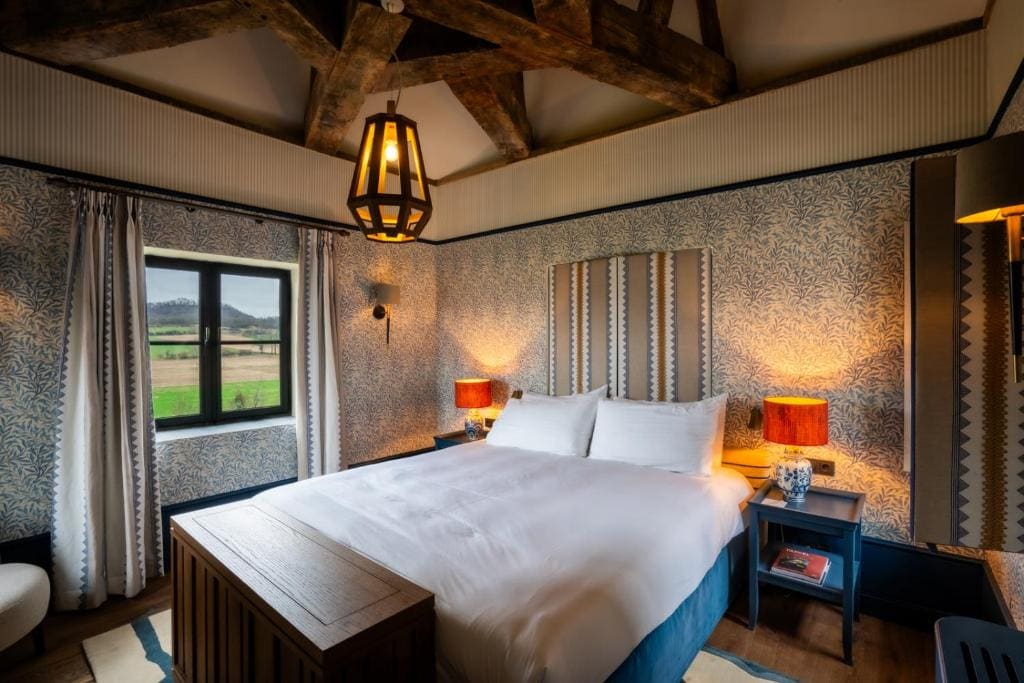 Cozy bedroom with a large bed, striped headboard, two patterned lamps on bedside tables, decorative wallpaper, exposed wooden beams, and a window showing a scenic countryside view.