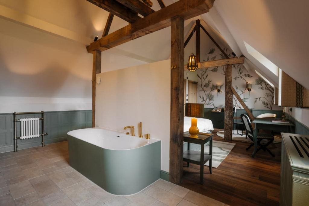 A spacious, rustic-style bedroom features exposed wooden beams, a freestanding green bathtub, a bed with patterned wallpaper behind, a desk area, and warm ambient lighting. The space combines modern and vintage decor.