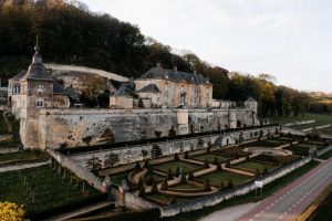 A large historic stone chateau sits atop a hill with manicured gardens and topiary trees below, surrounded by stone walls and lush greenery, beside a quiet road.