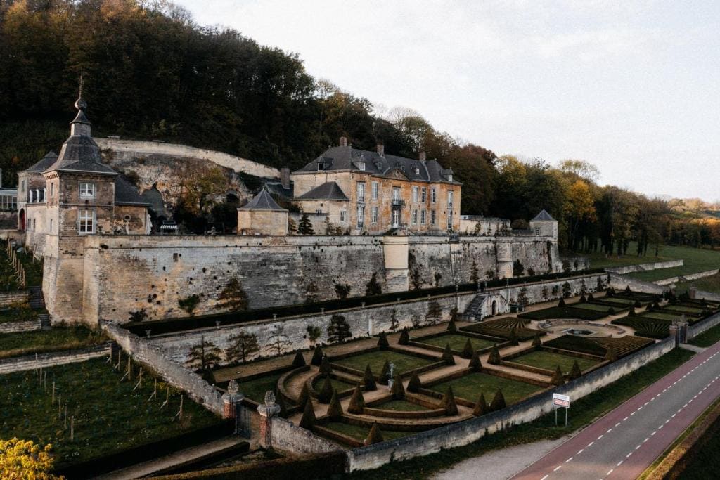 A large historic stone chateau sits atop a hill with manicured gardens and topiary trees below, surrounded by stone walls and lush greenery, beside a quiet road.