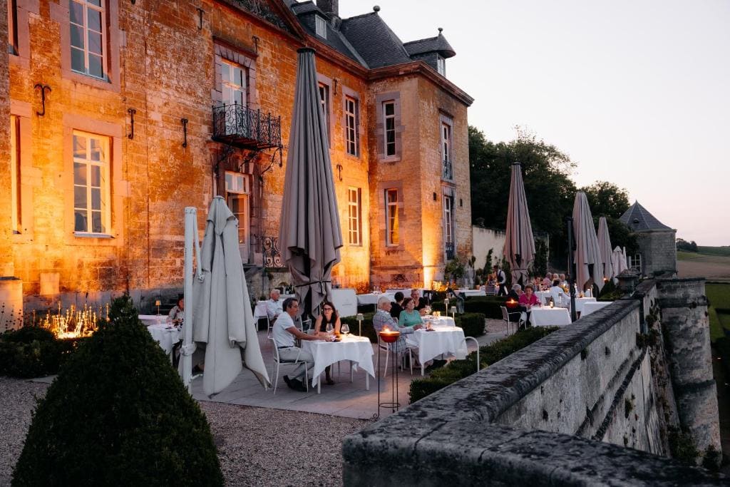 People dine at elegant outdoor tables beside a historic stone building at sunset. Umbrellas, greenery, and soft lighting create a cozy ambiance in the courtyard of what appears to be a grand, old mansion or chateau.