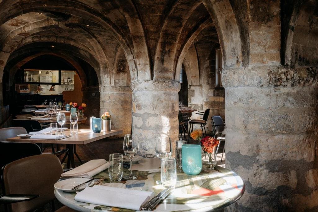 A cozy restaurant with stone arches and pillars, set with round and rectangular tables, glasses, napkins, and small vases of flowers; warm light creates an inviting atmosphere.