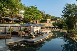 Luxurious resort pool area with modern lounge chairs, cabanas, and umbrellas surrounded by lush greenery and multi-story buildings under a clear blue sky. The serene setting reflects in the calm water.