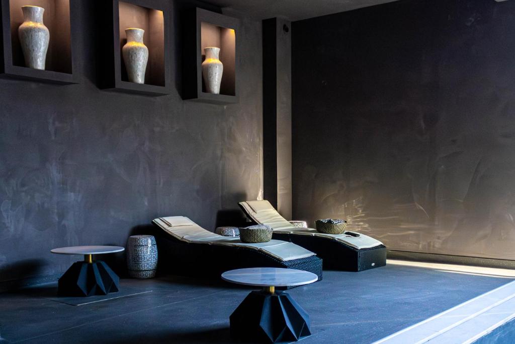 A modern, dimly lit lounge with two reclined chairs, two round tables, and decorative vases displayed in wall niches above. The walls and floor are dark, creating a calm, minimalist atmosphere.